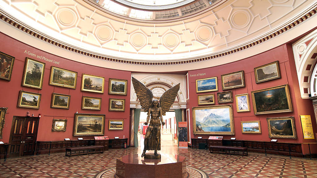 A bronze statue of an angel holding out her hands is central to a round room with lots of oil paintings on the walls. The beauty of the room radiates from the photo, capturing the time before the halls of the museum are filled with visitors