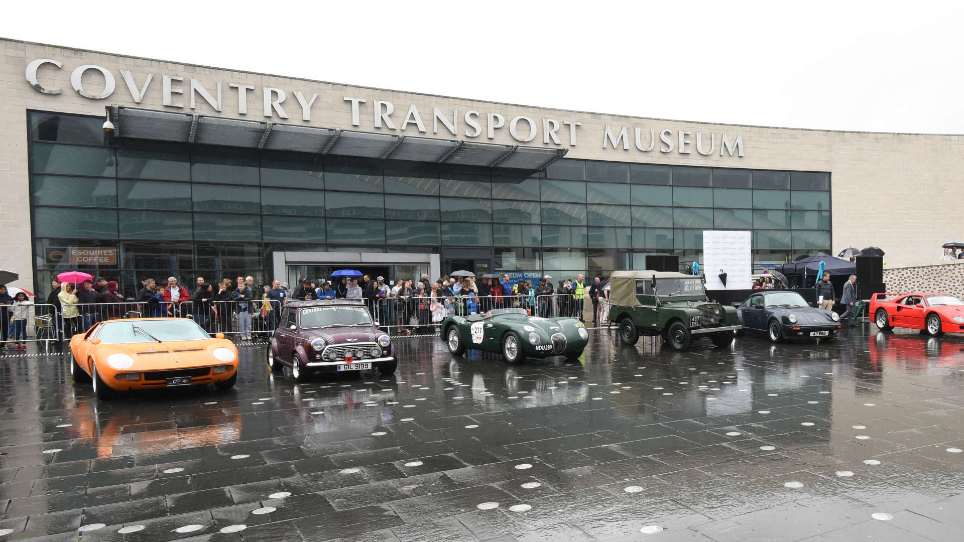 A variety of cars, from classic to very modern sleek vehicles, are parked in front of a large building with a glass front. You can tell it's a gloomy day from the rain on the ground