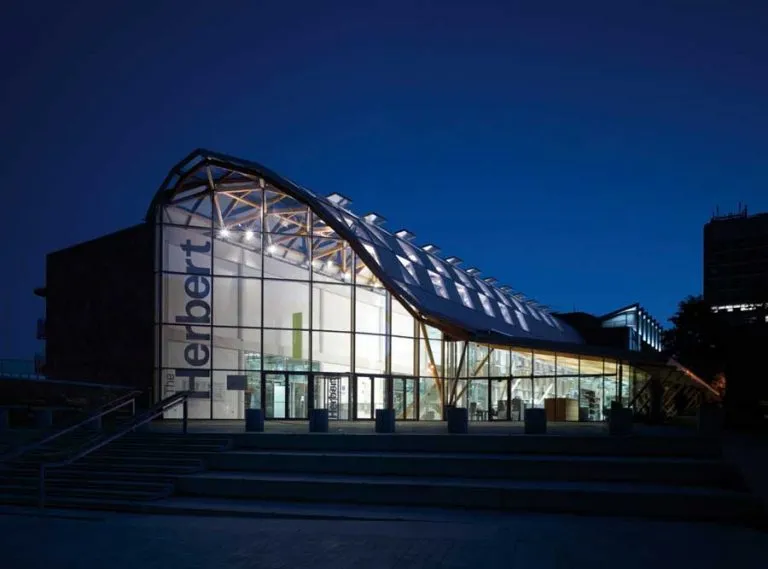 On a dark night, the impressive and tall Herbert museum is lit up and dazzles the night sky. The glass panes that cover the front let light out into the night