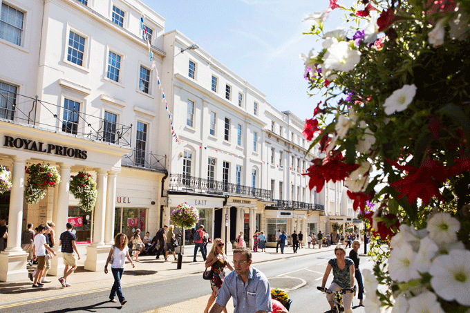 The white façade of the shops on the street is clean and bright, reflecting the summer sun. The architecture appears to be in a classic style that harks back to the Victorian era. People walk happily across the street towards the shops. In the foreground of the picture is a bush with pink and white flowers in complete bloom, making the picture have a feeling of freshness and new life.