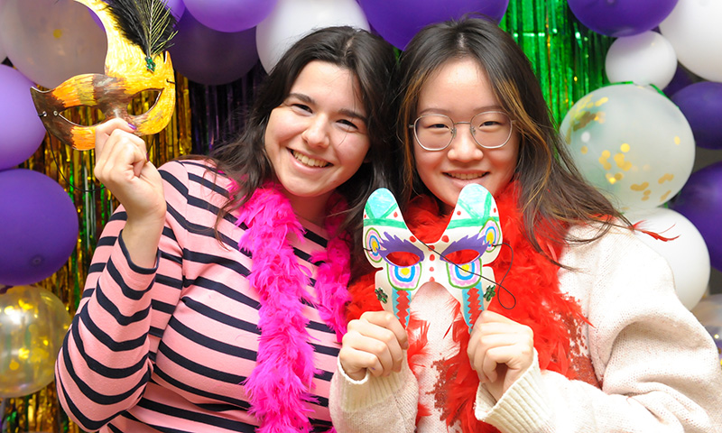 Two students smile at the camera, holding mardi gras masks against a balloon backdrop. 