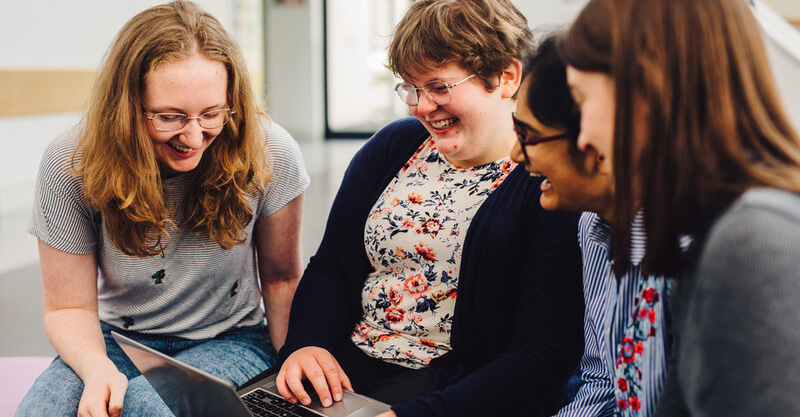 Four students sit together around one laptop, laughing at the screen.