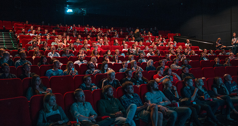 A group of people sit in a darkened movie theatre, looking towards the screen.