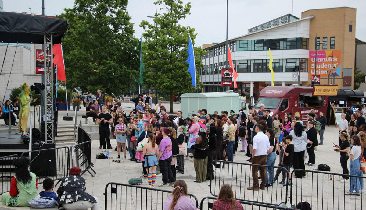 A stage outside on the PIazza with a performer on stage and a crowd of people watching, festival flags can be seen in the background and the SU building in the distance