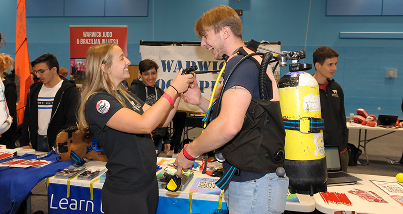 A student exec demonstrates how to use scuba diving equipment to a laughing first year. 