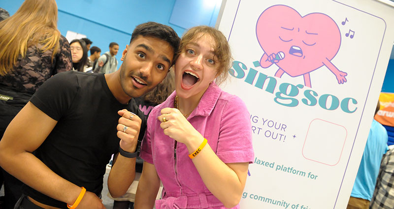 Two students from Sing Soc smile as they hold pretend microphones