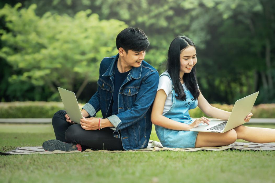 Two Asian students sitting on the grass back to back holding laptop in their hands 
