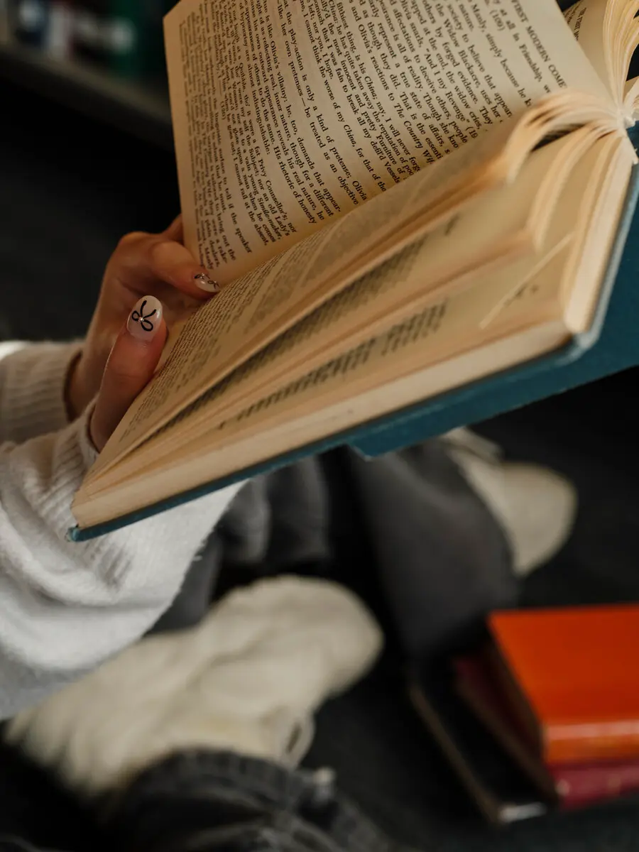 A student reads a book whilst sat on the floor beside a small stack of textbooks.