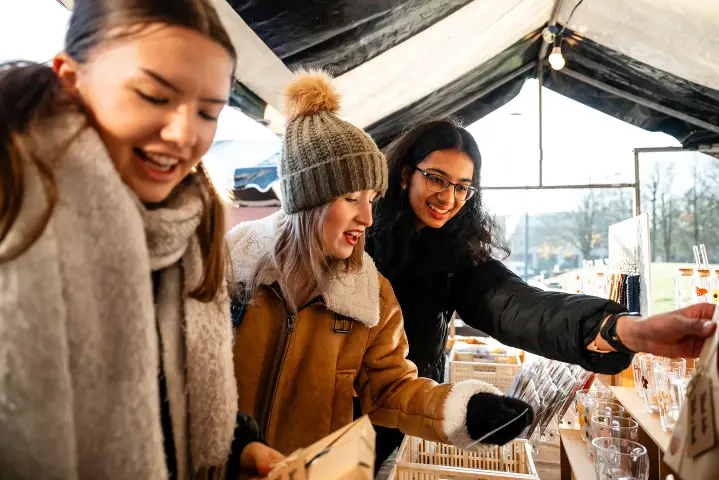 Three students are looking at items for sale at an outdoor craft market.