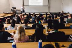 A lecture theatre with around 50 students sitting in rows. There is an academic at the front talking in front of a powerpoint