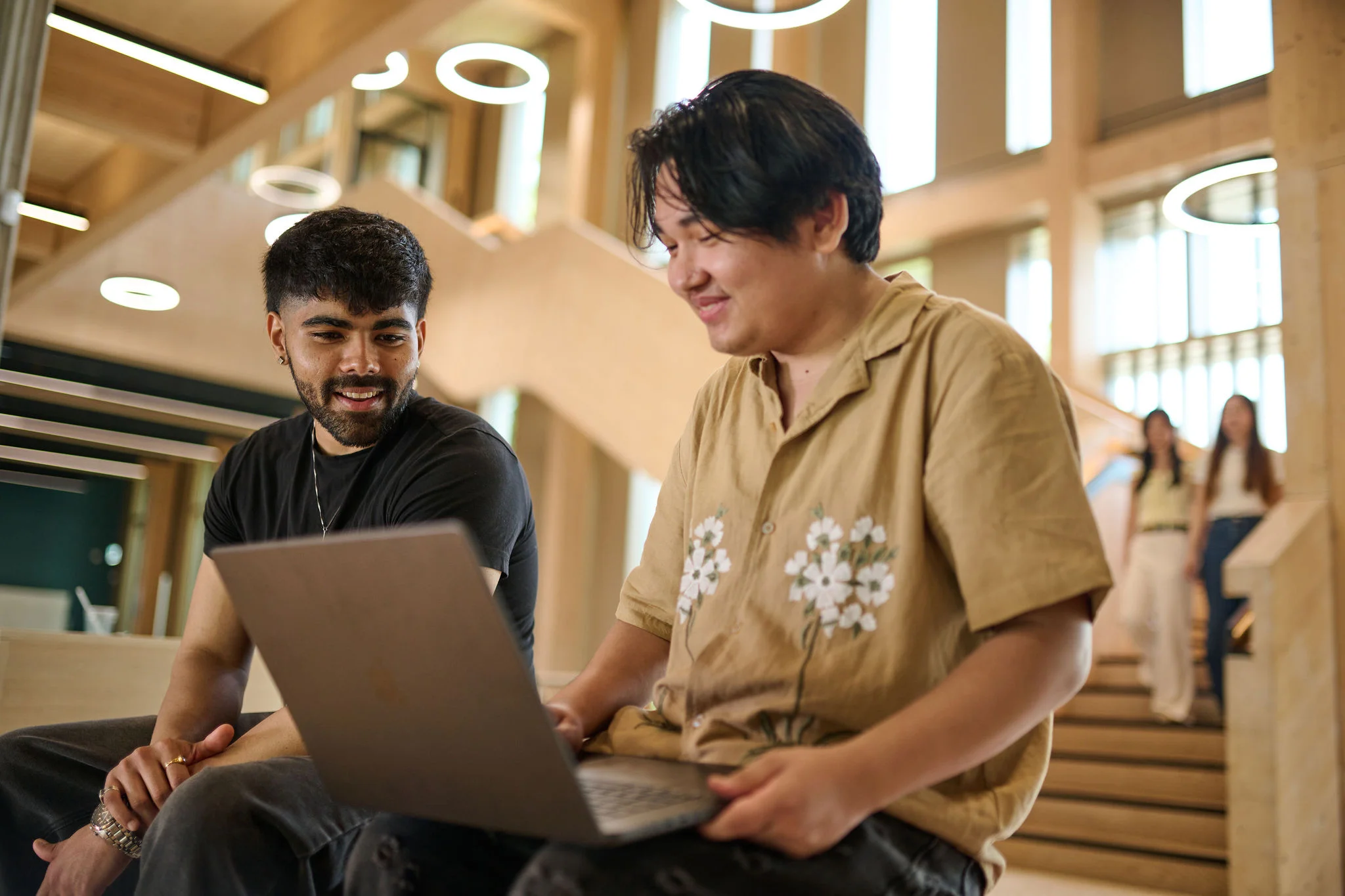 Two students are sitting in a brightly lit campus building looking at a laptop screen.