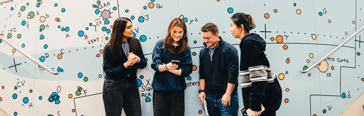 Four students engaging in discussion beside a mural illustrating global themes.