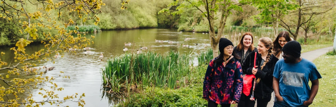 Students walking along a tree-lined river at the University of Warwick.