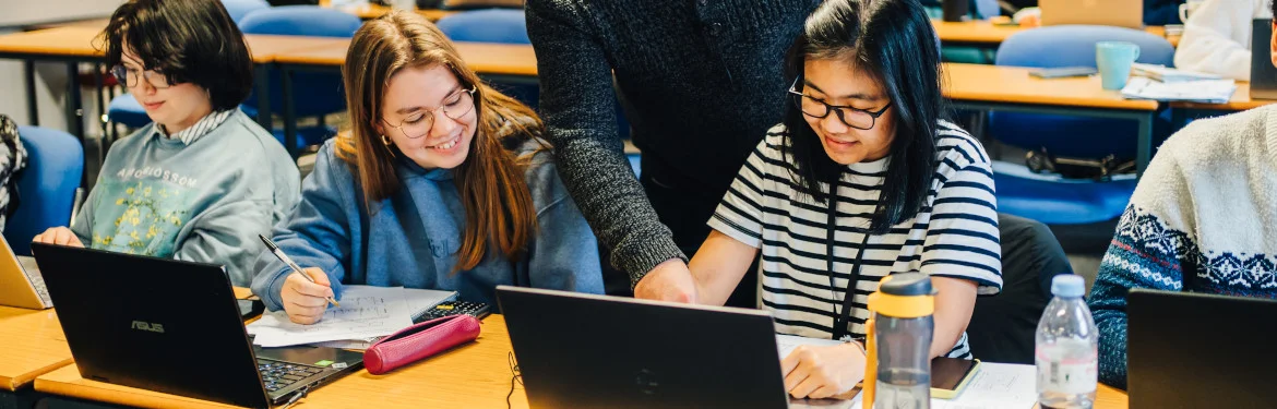 Students work on laptops in a classroom while an instructor provides guidance.