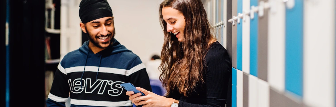 Two students smiling and looking at a phone in a corridor