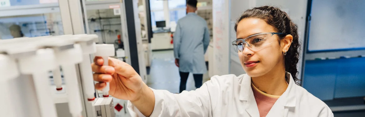 Student in a lab coat inserting a sample into laboratory testing equipment.