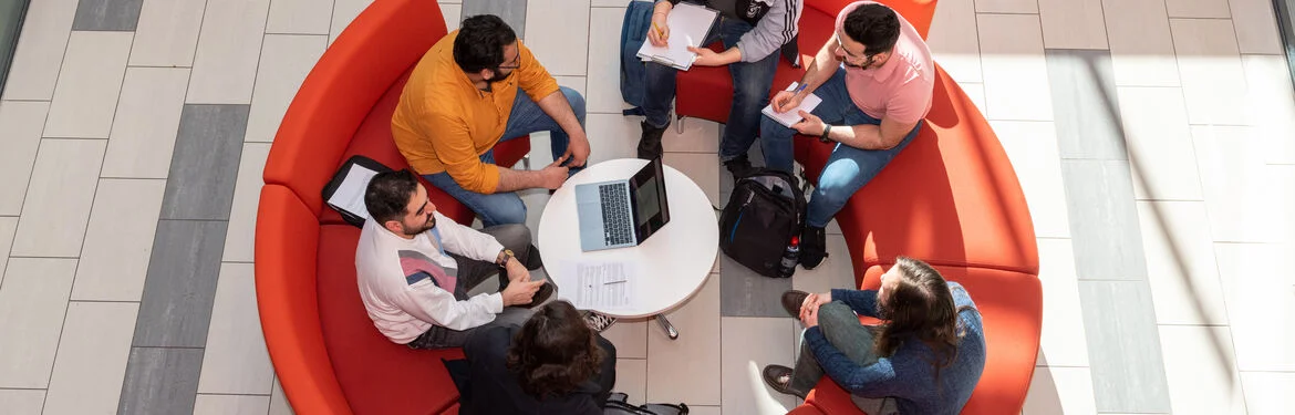Students gathered around a table on a curved orange sofa, collaborating with a laptop and notes.
