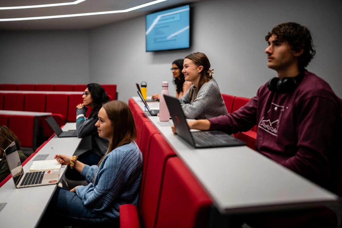 Students sitting in a lecture theatre with laptops on the desks in front of them.
