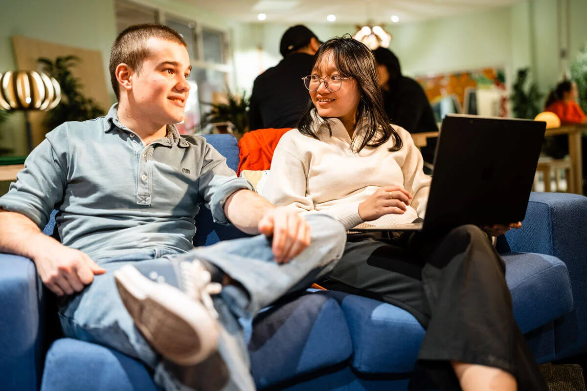 Two people sat on a sofa in conversation, one with a laptop open in front of them.
