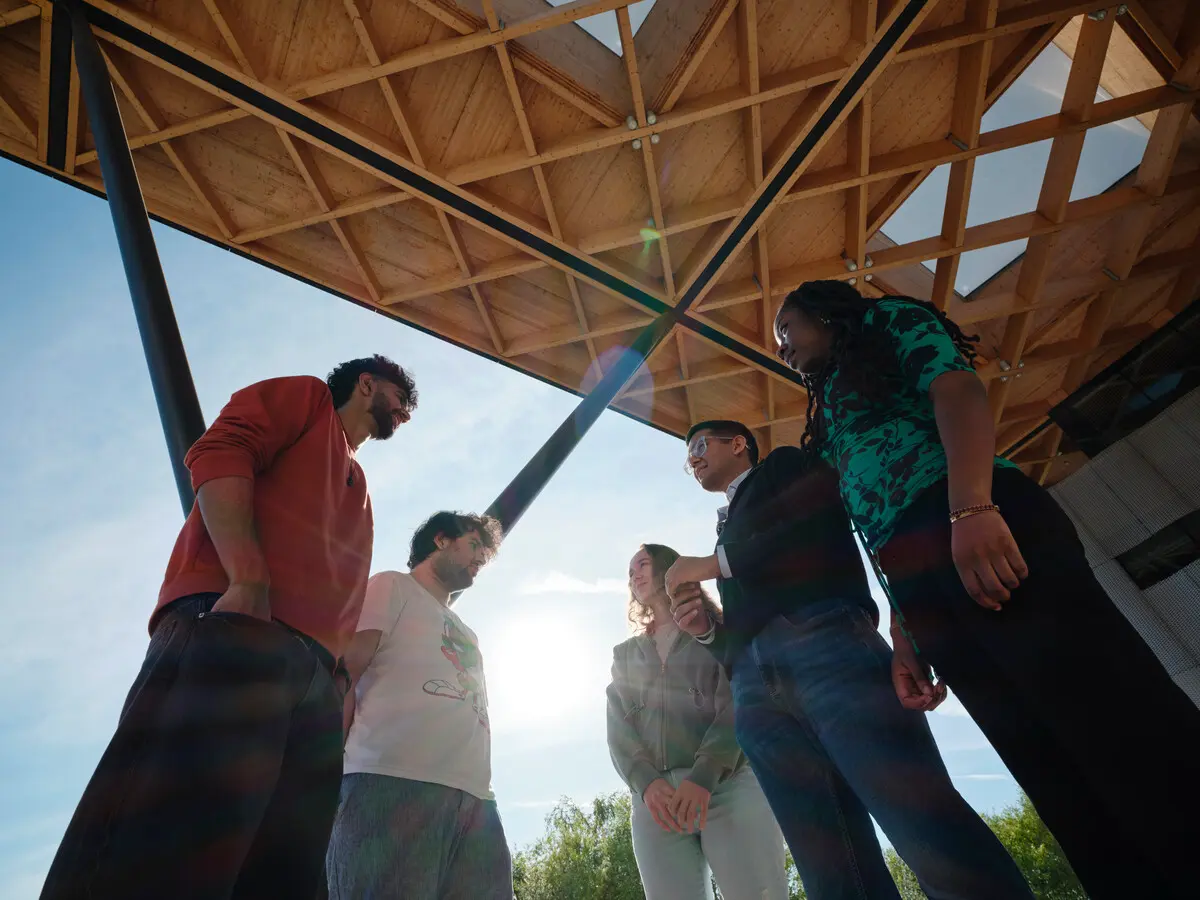 Five students stand beneath a wooden roof with prominent crisscrossed beams, talking together on a bright sunny day.