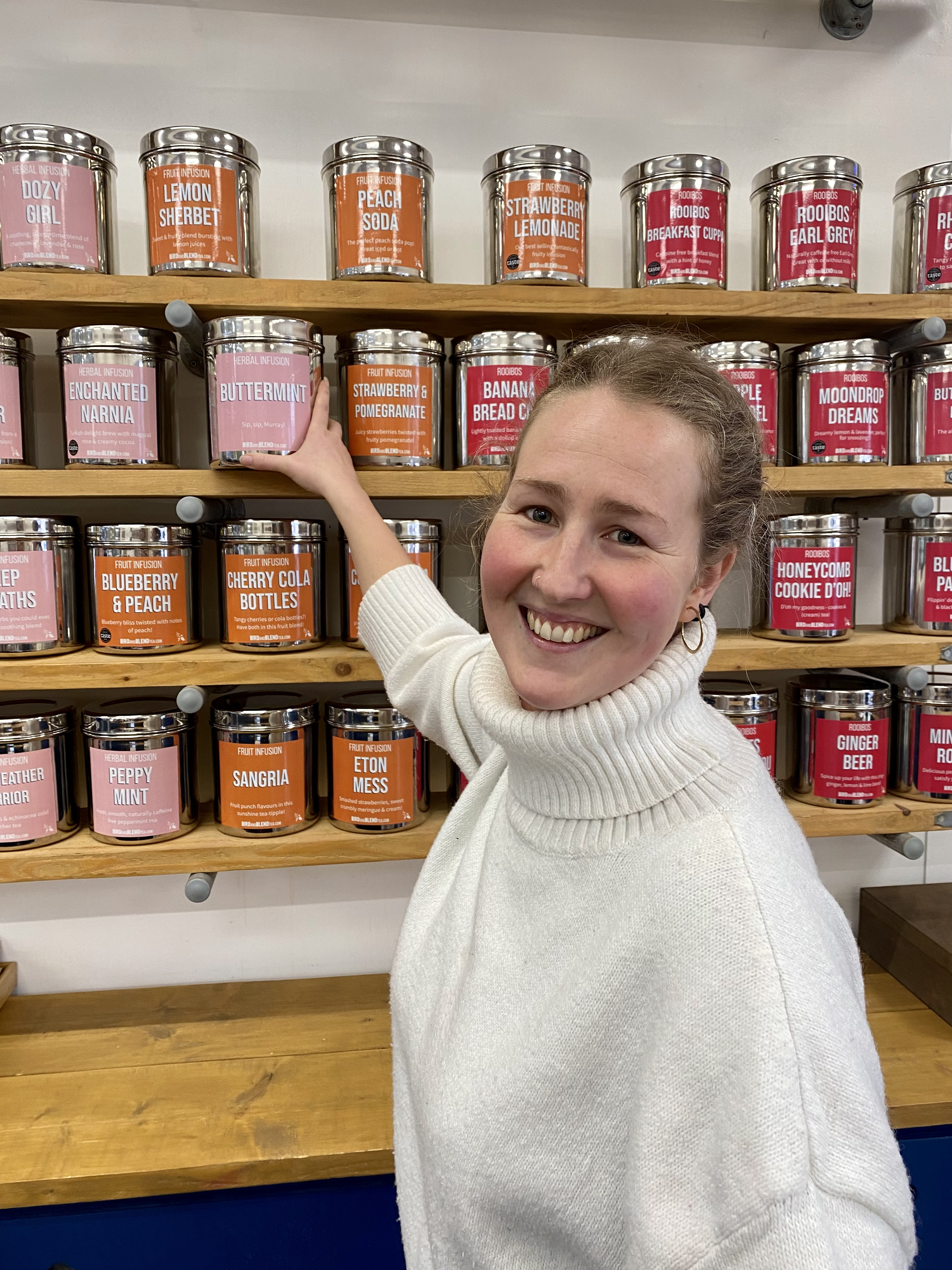 Image of women smiling in a white turtle neck jumper, hair tied up and wearing gold hooped earrings - reaching for a tin of tea on wooden shelves full of tins