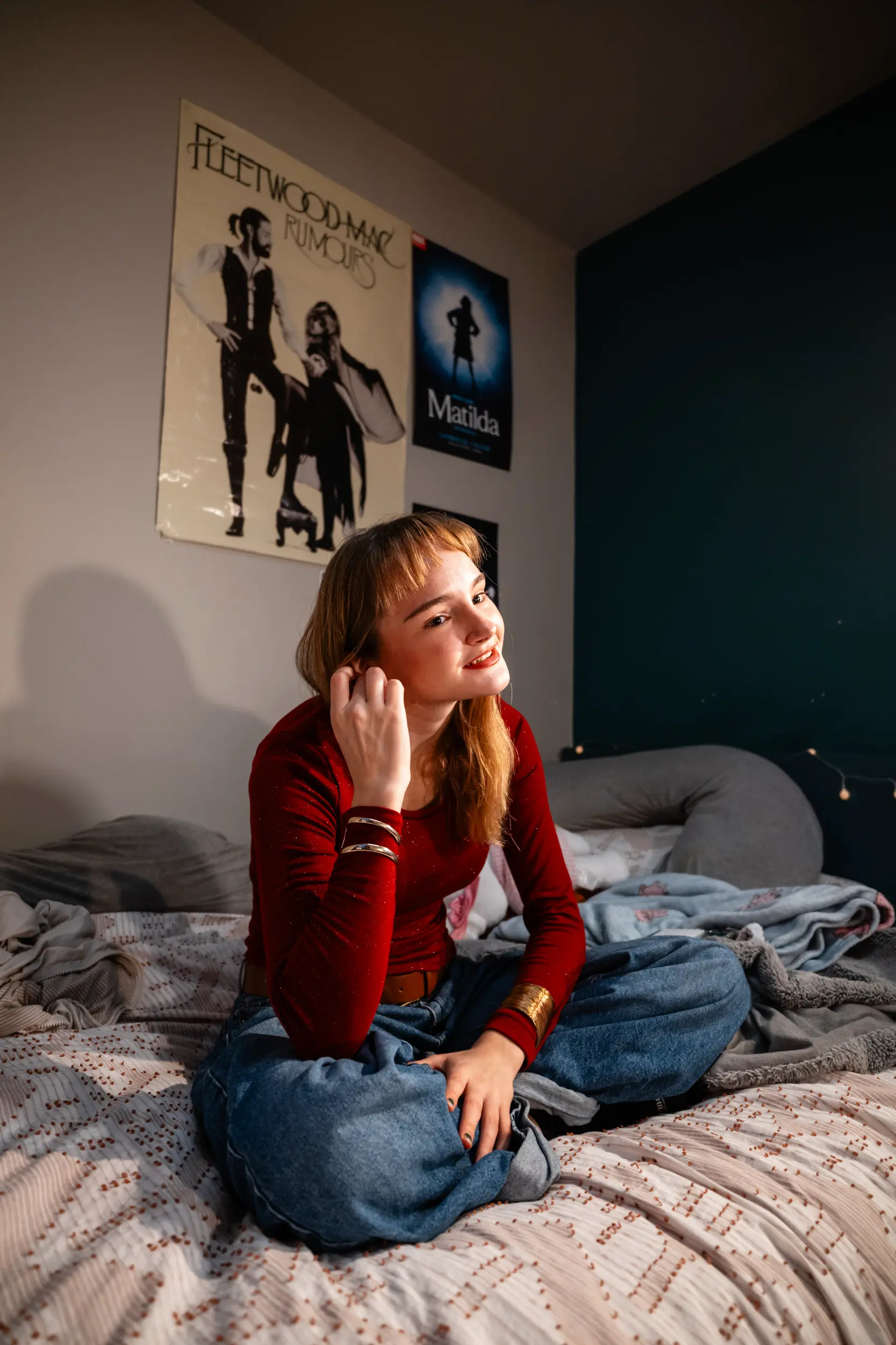A student in a red top sits on a bed in university accommodation with theatre posters on the wall behind them.
