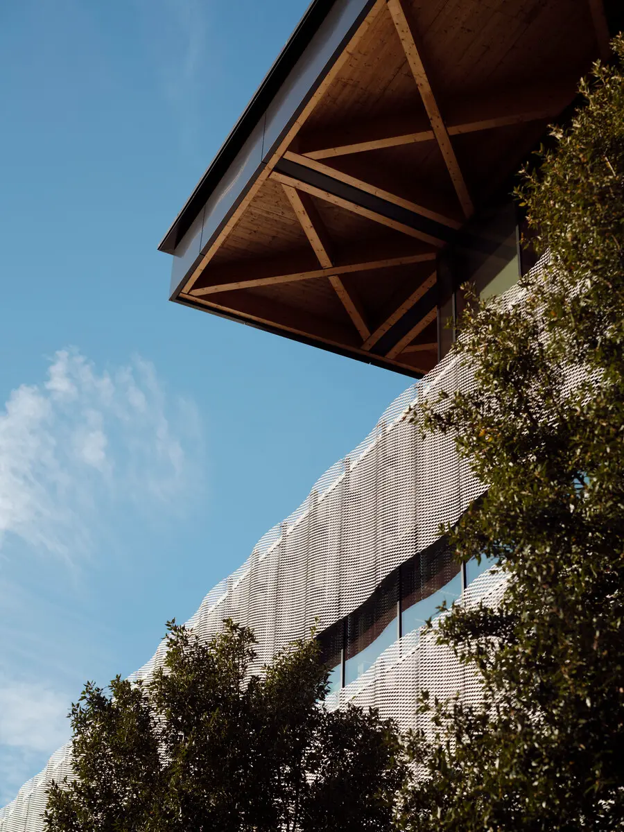 Low-angle view of a modern building with a textured, wavy facade and large windows, partially obscured by leafy trees, under a clear blue sky.