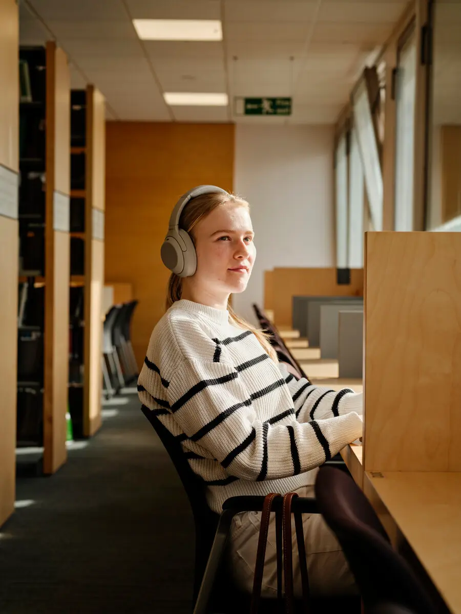 A student with headphones on sitting in a shared study space staring into the distance.