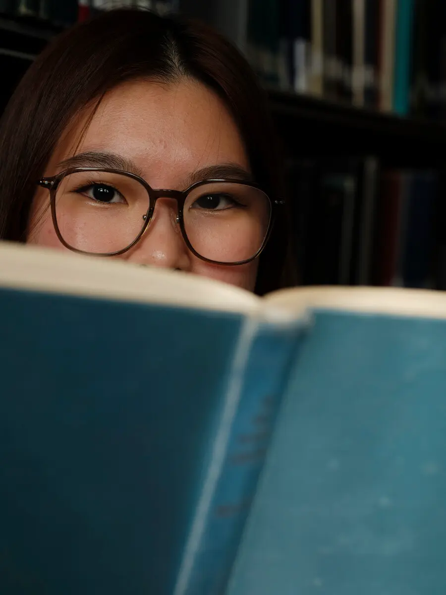 A student is looking at the camera over the top of an open book.