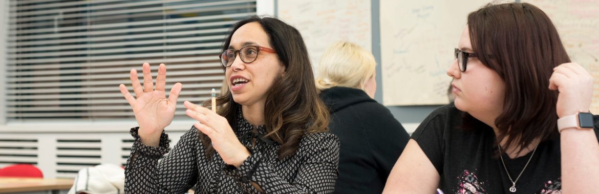 Two female students in a classroom setting. One of them is speaking passionately and the other one listening