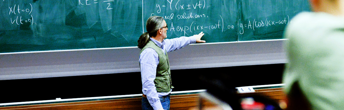 This image shows a university professor point out a maths equation on a chalkboard to students in a lecture theatre