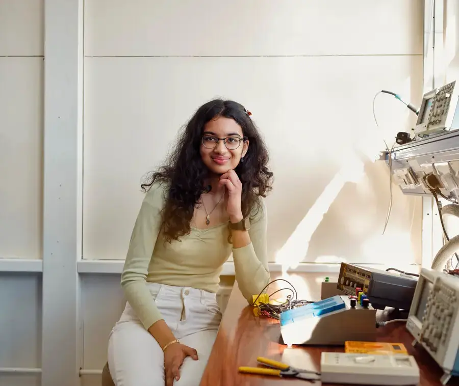 A student sits at a desk with electronic equipment, smiling casually at the camera.