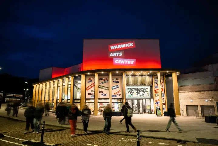 The Warwick Arts Centre building lit up at night with people milling around in front of it.