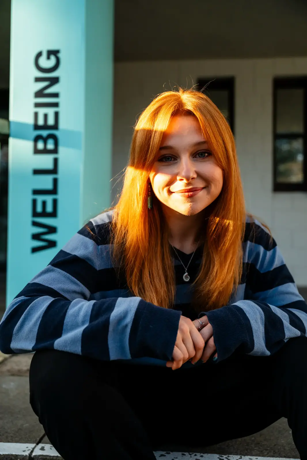 A student in a blue stripy jumper sits in front of a wellbeing sign, smiling at the camera.