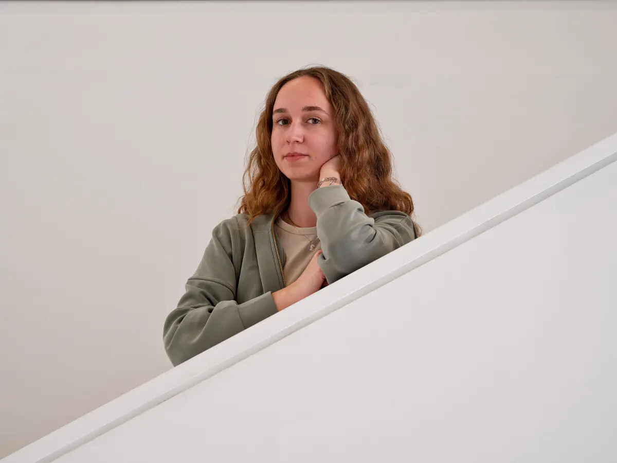 A university student leaning on a staircase railing indoors, looking at the camera.