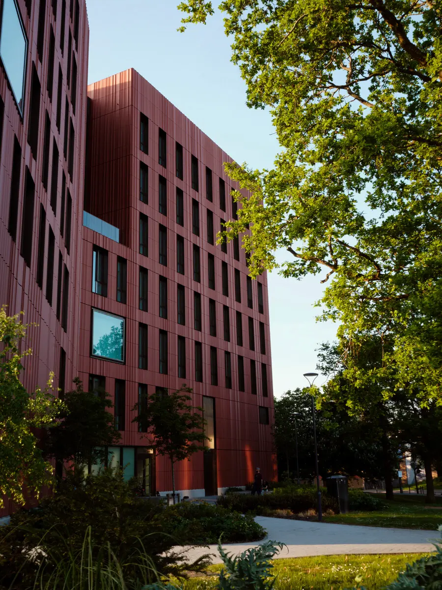 A tall red campus building with lots of windows, surrounded by trees.