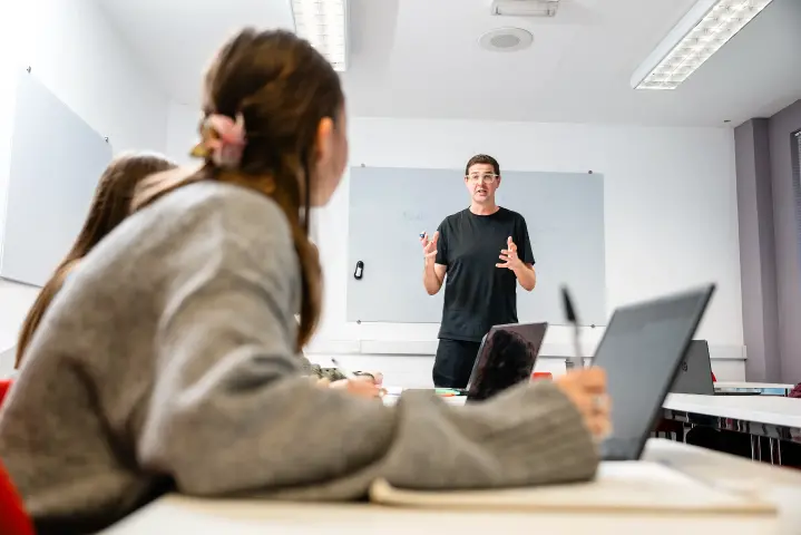 A university lecturer explains materials on a whiteboard while students in the foreground take notes on laptops and in notebooks.