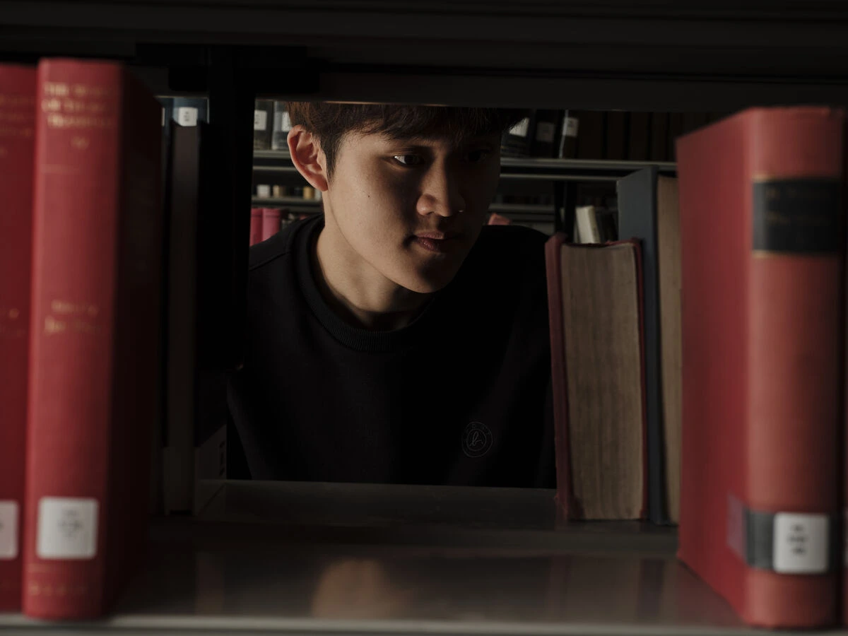 A student in the library looking through a gap in a shelf of books.