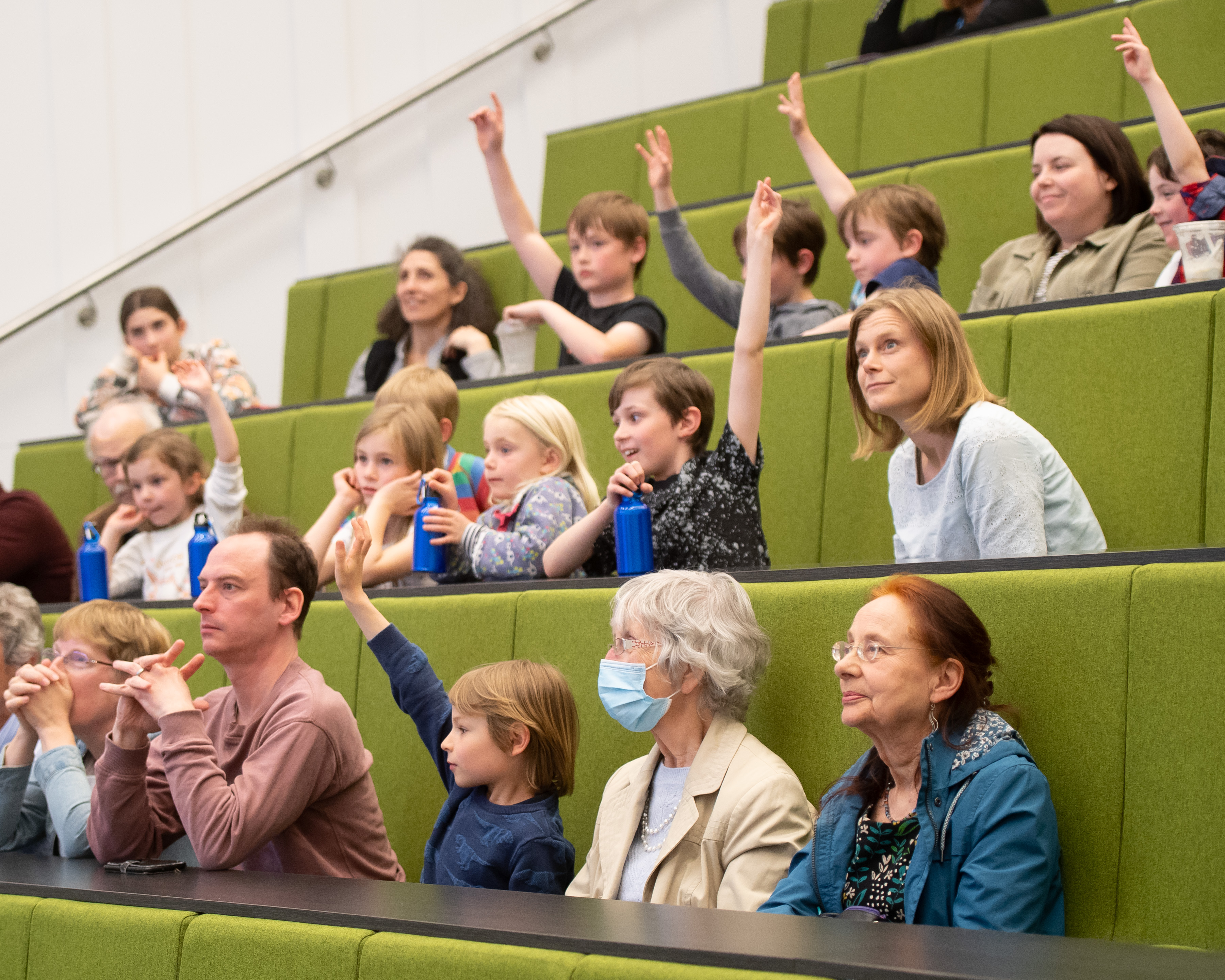 An audience of children raise their hands to answer a question
