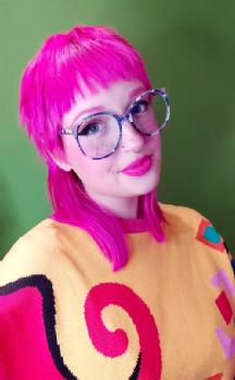 Edie Jo Murray - This image shows Edie, who has long pink hair, glasses, and a yellow patterned jumper, against a green background.