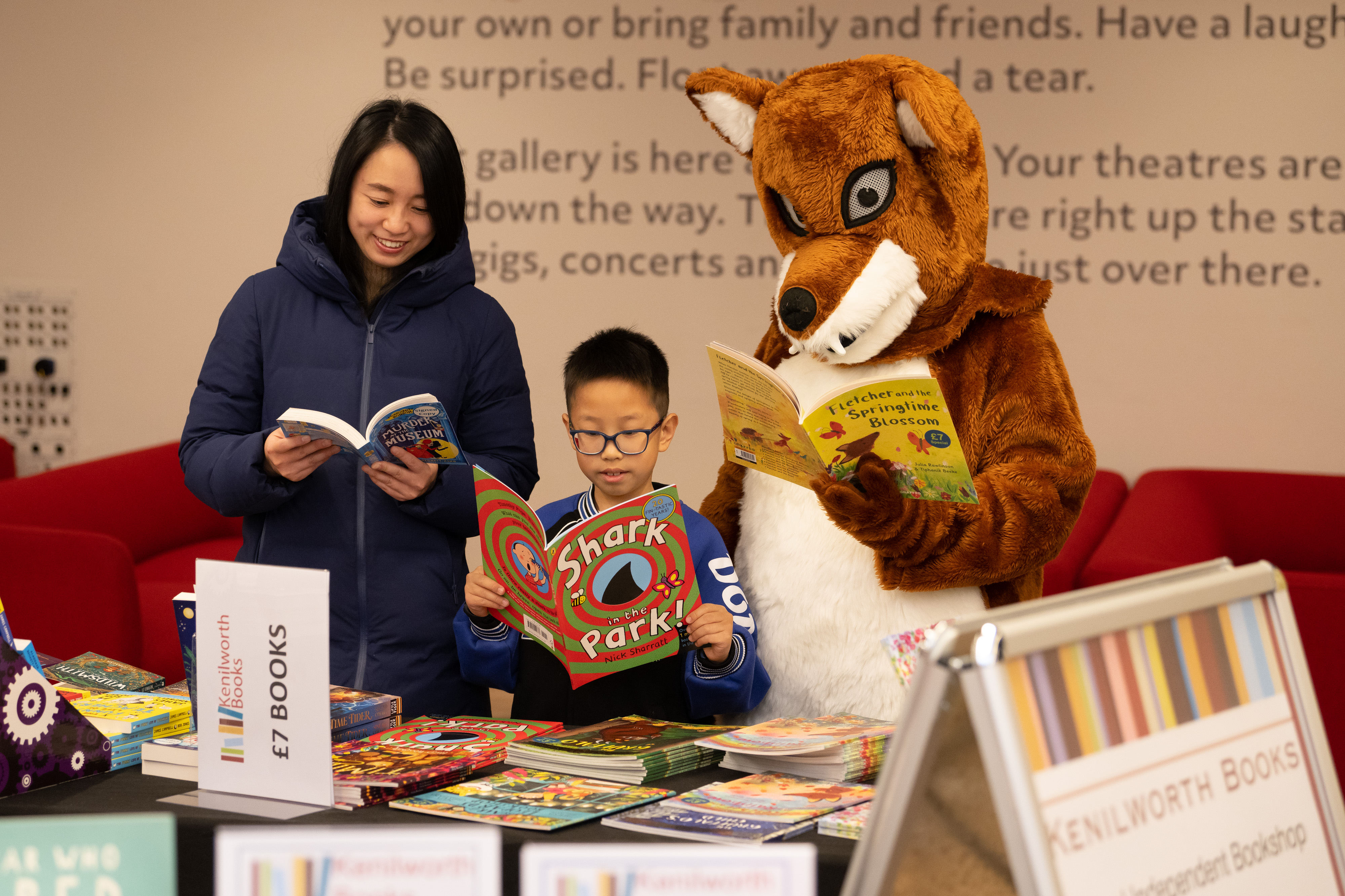 A woman and child read books. Next to them is someone in a fox costume. 