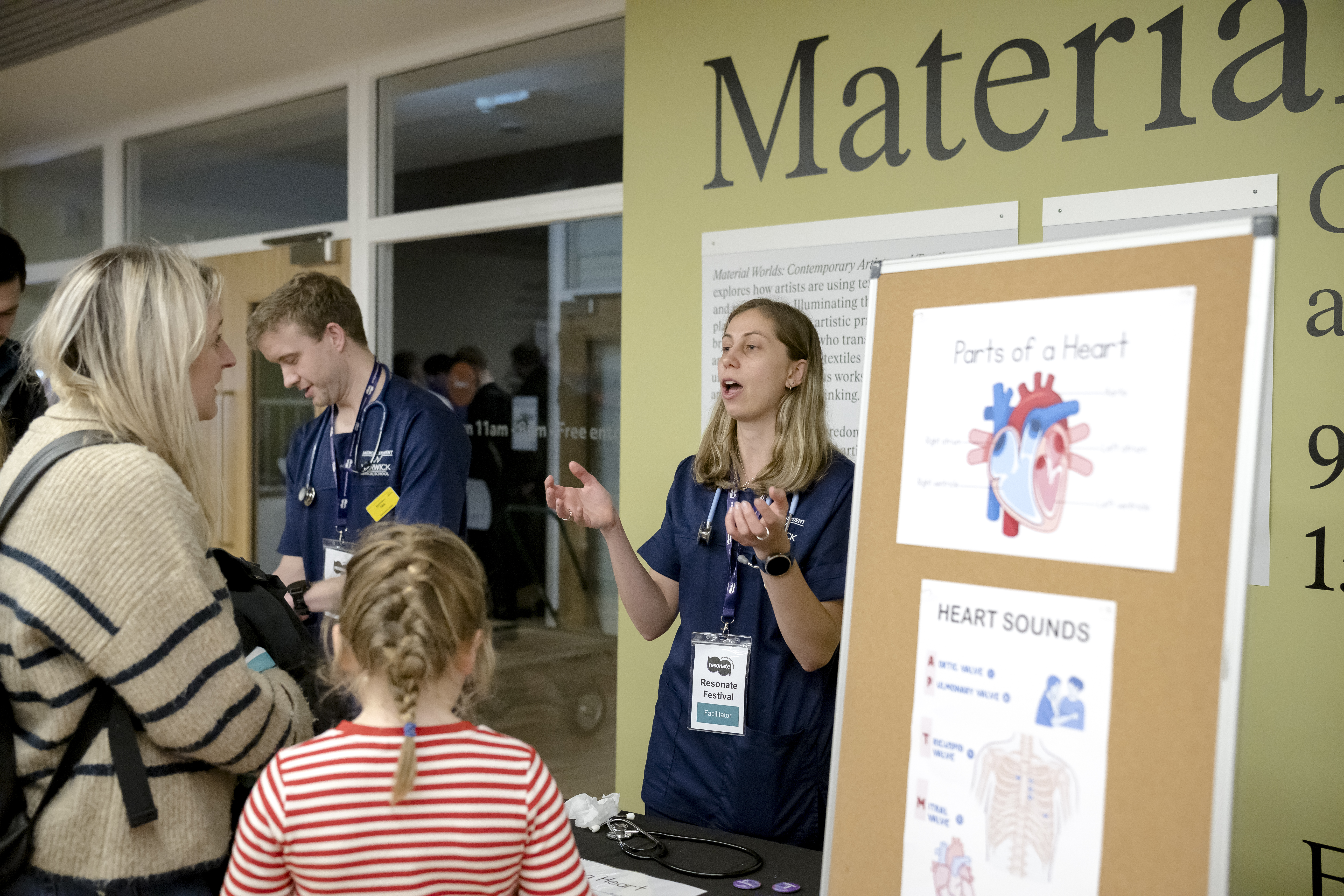 A researcher stood behind a table and explaining their research to attendees. The poster by the researcher shows a diagram of a heart. 