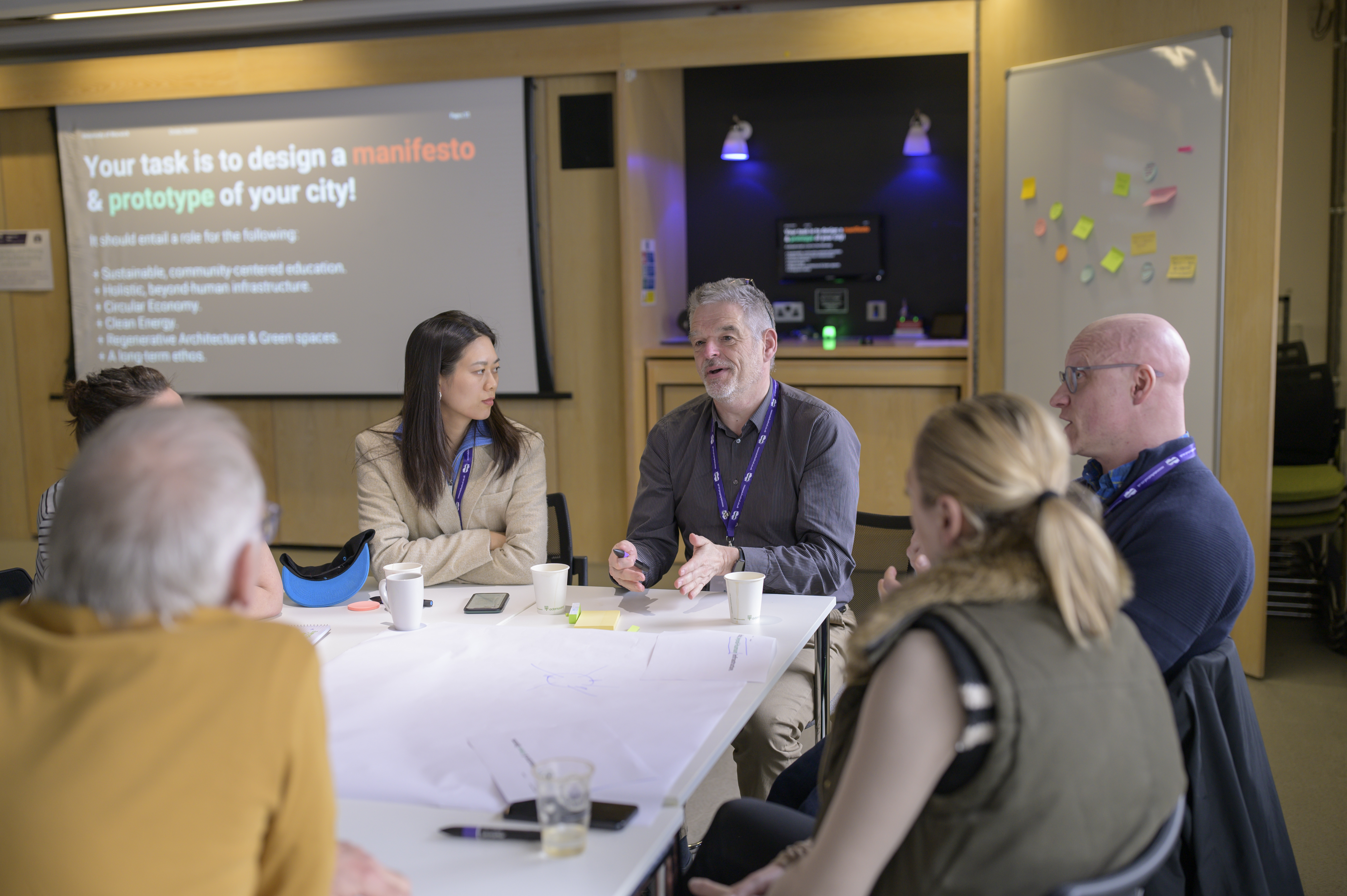 A group are sat around a table, one person is talking and others listen. 