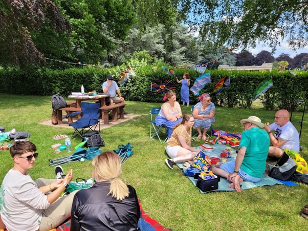 Groups of people sitting on grass, picnic chairs and benches, chatting and eating food together in a green space with grass, a hedge and trees. There are various types of pride flags in the background, in different colours.