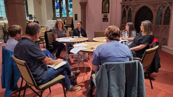 A group of people sitting in a circle, with some papers on tables. They are all looking at the person who is speaking. 