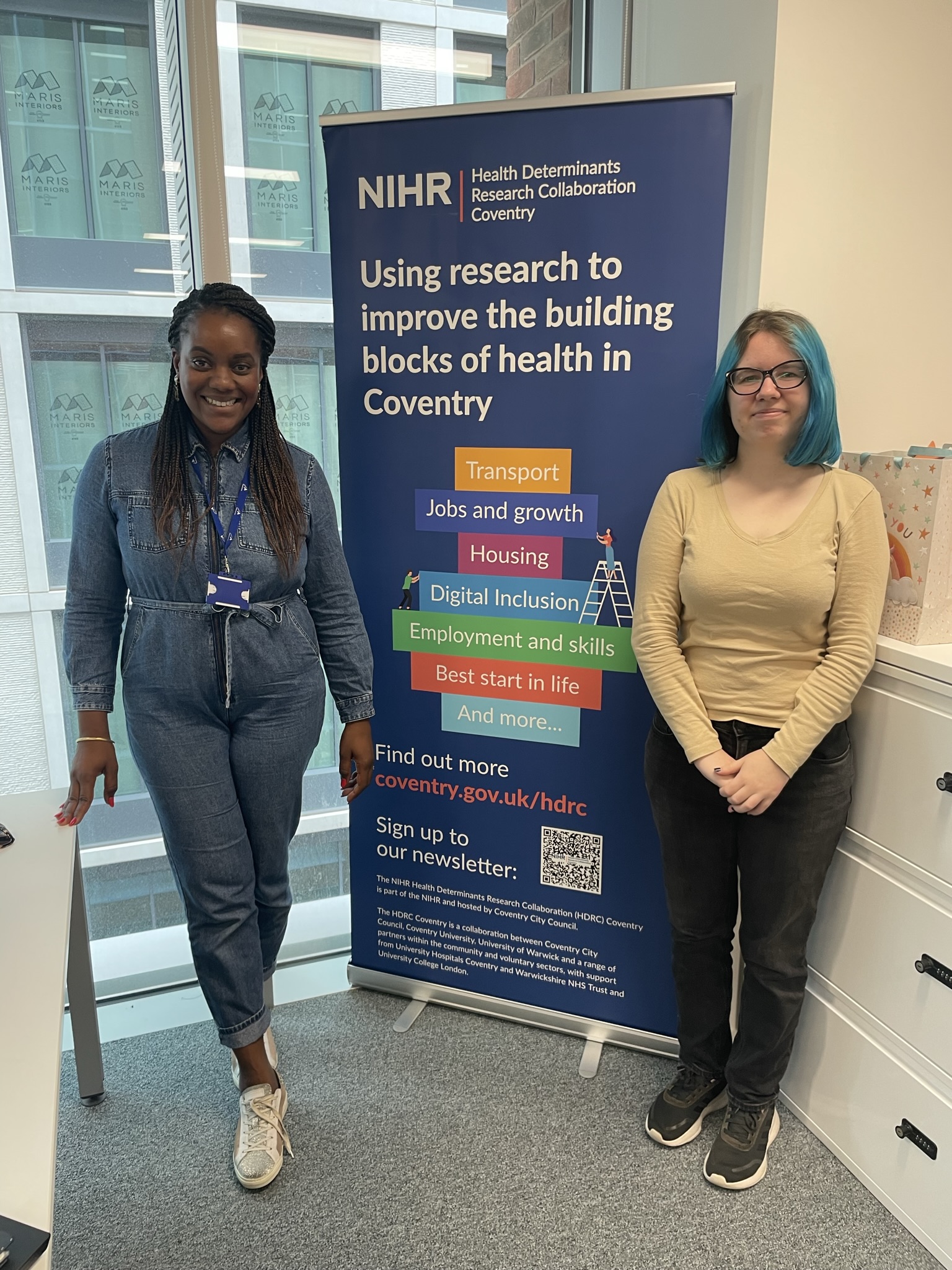 Jessica Weeks and Ida Slavina stand in front of a blue sign that says 'NIHR - using research to improve the building blocks of health in Coventry'