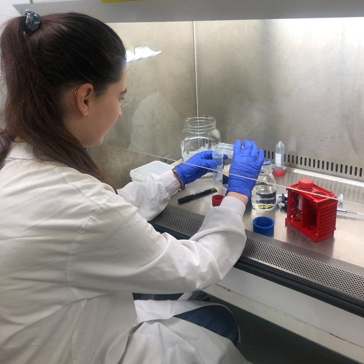Mimi is sitting at a fume cupboard, wearing a lab coat and blue gloves. She is pouring from one vessel into another. 