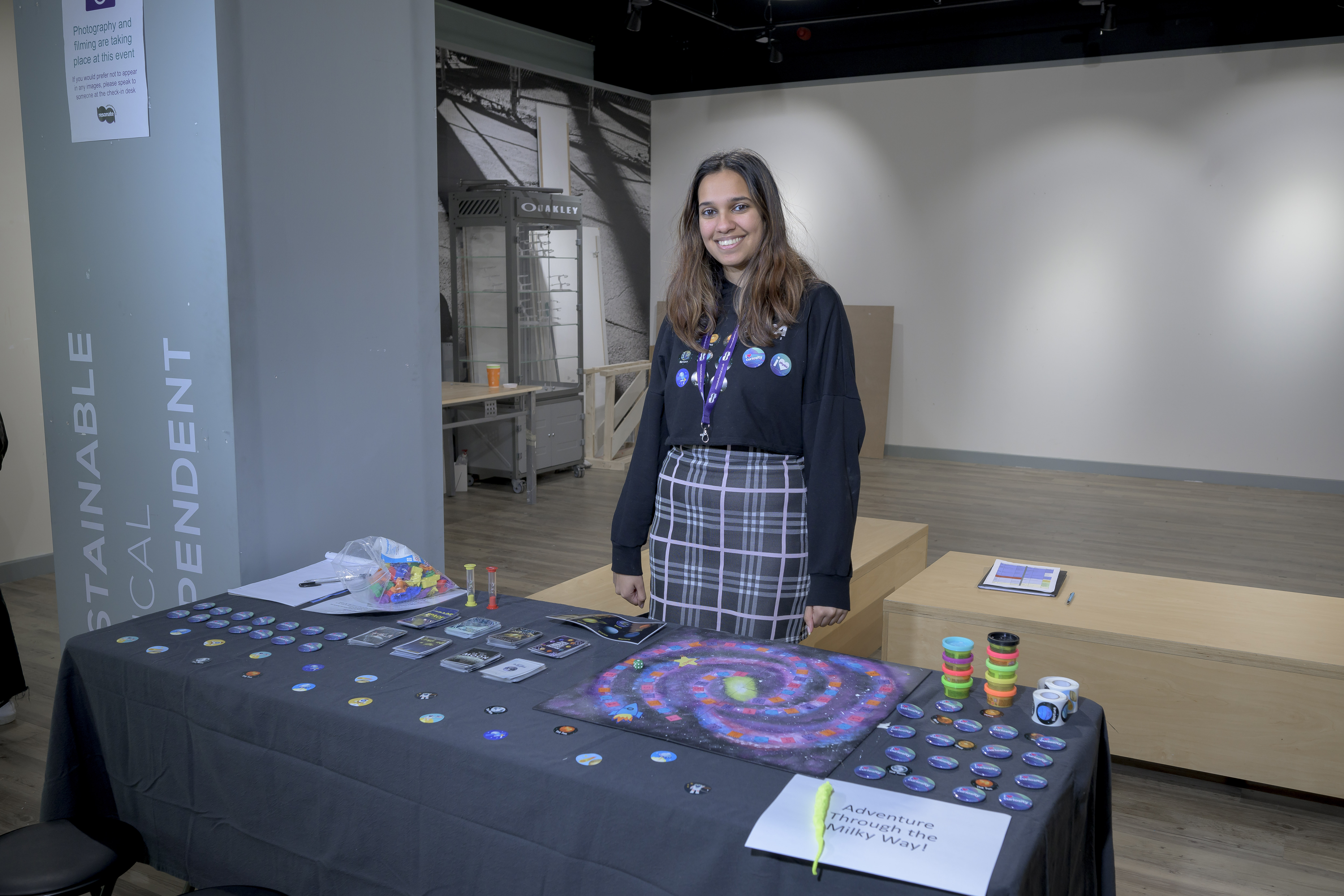 Gillian is standing behind a table with a black tablecloth on it. On the table is a board game painted in a swirl of purple and blue. On the table are small 3D transport-shaped objects to use as counters. There are also cards the size of playing cards, modelling clay or play dough, stickers and badges.