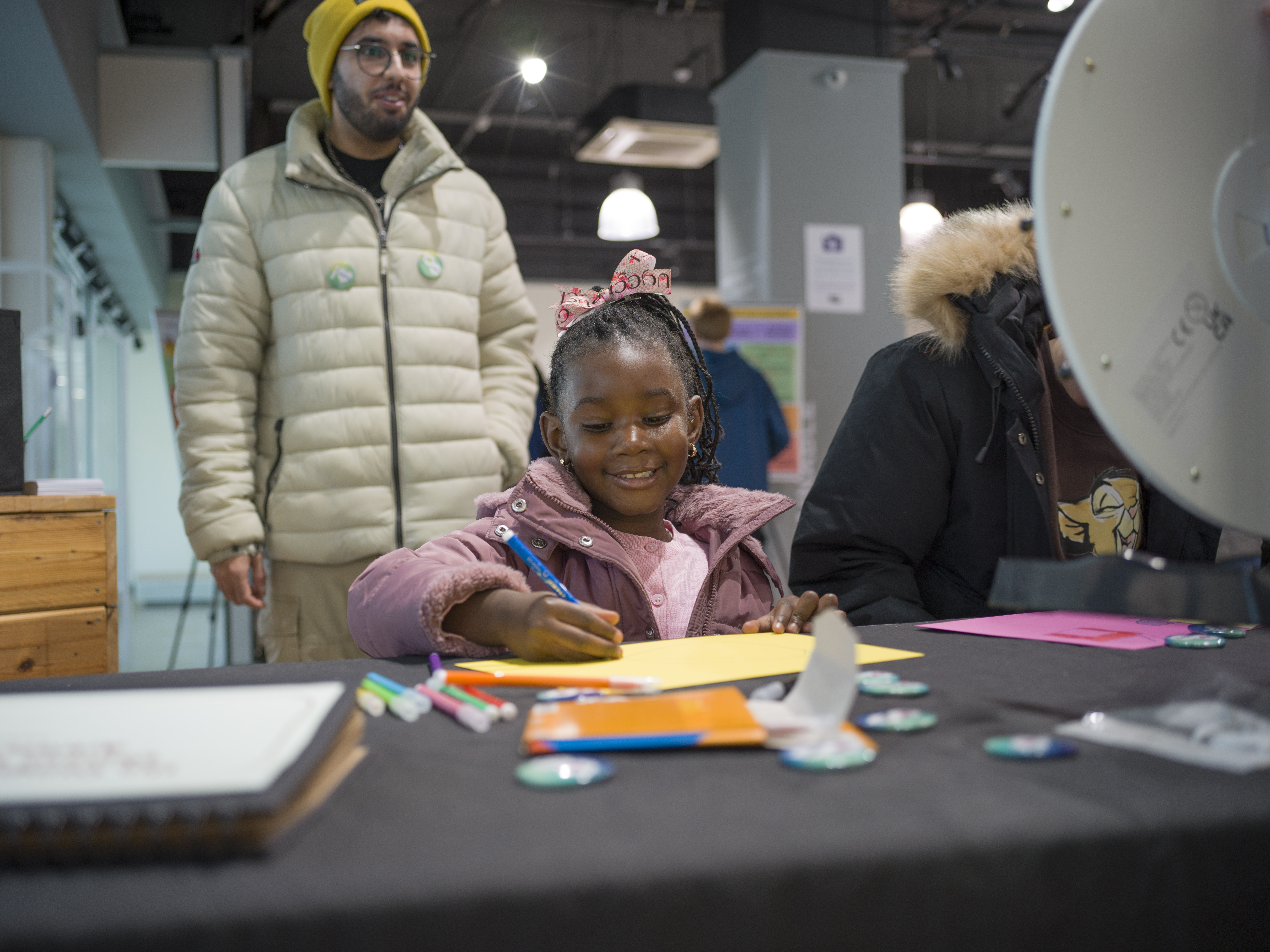 A child drawing on yellow paper. An adult watching in the background. 