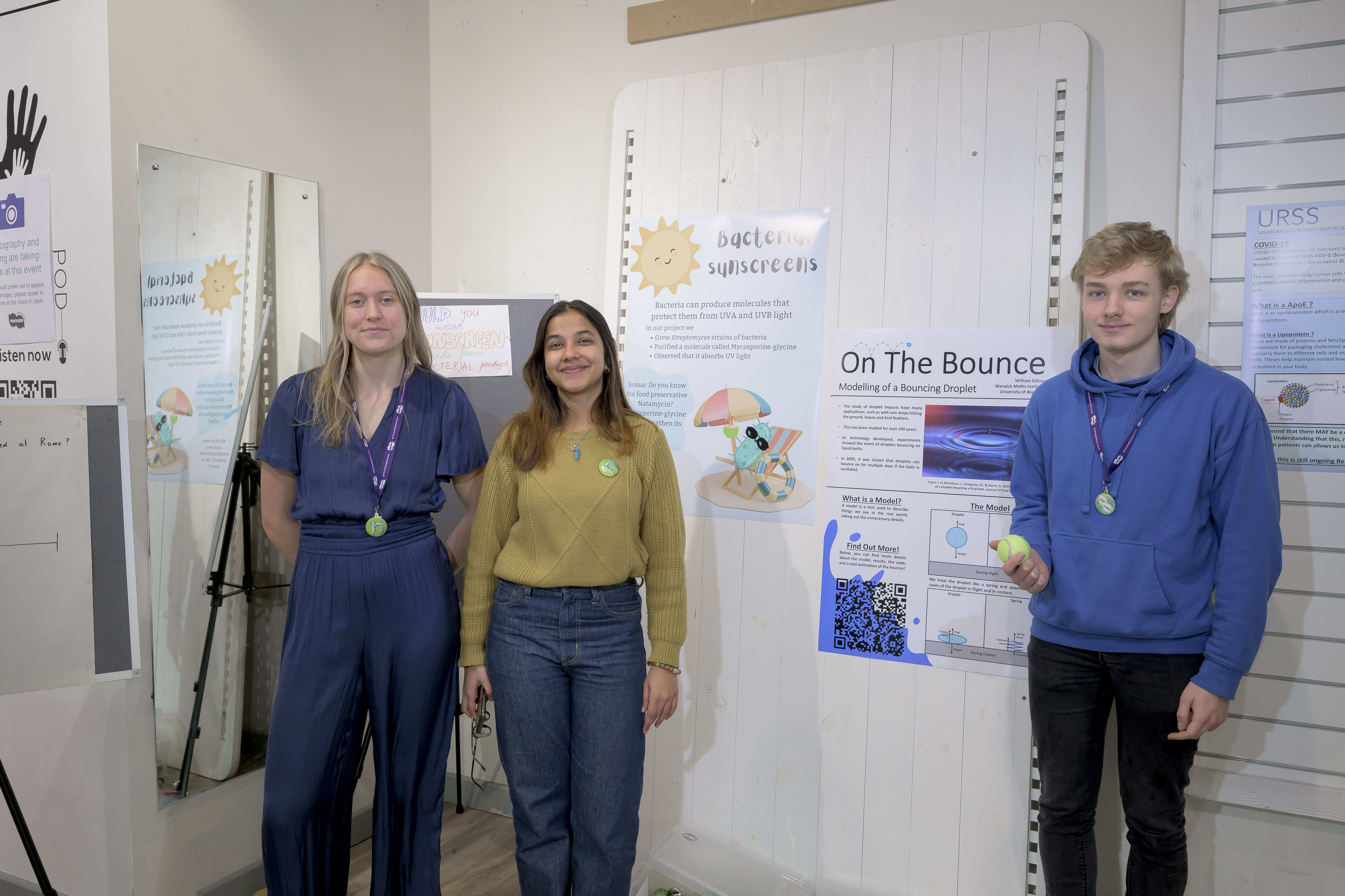 Three students standing with their posters. One of the students is holding a tennis ball and their poster is titled 'on the bounce'. 